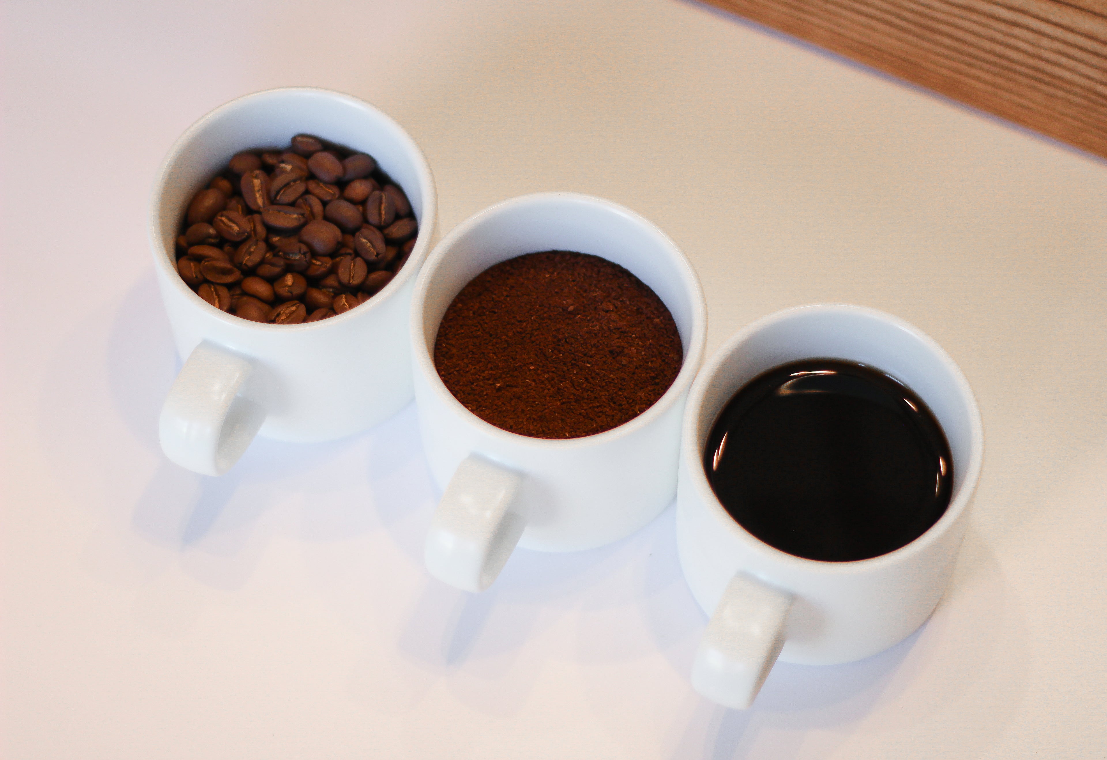 3 white mugs filled with whole coffee beans, ground coffee beans, and brewed coffee